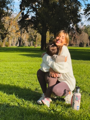 Mujer sonriente de pelo rubio abraza a una niña en un parque. Ambas visten suéteres claros y pantalones oscuros. Hay un termo en el suelo