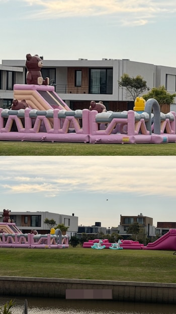 Estructura inflable rosa y marrón con toboganes y figuras de osos sobre césped verde, con casas modernas al fondo y un canal de agua al frente
