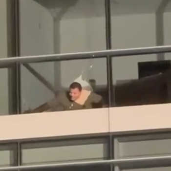 Un hombre con camisa verde mira a través de una ventana con el cristal fracturado. Fragmentos de vidrio roto rodean su rostro