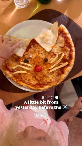 Una pizza con forma de cara de gato, decorada con aceitunas, un tomate y papas fritas, junto a la mano de una niña pequeña