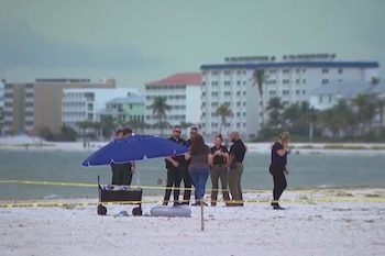 Una playa con arena, agua del mar, edificios al fondo, palmeras, varias personas, algunos agentes de seguridad, un paraguas azul, un carrito y una cinta amarilla acordonando el área