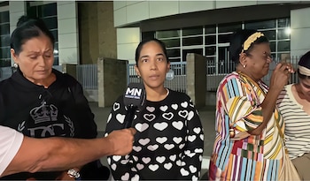 Primer plano de una mujer con suéter negro de corazones blancos hablando a un micrófono MN, flanqueada por dos mujeres. Una mira hacia abajo, la otra se ajusta gafas
