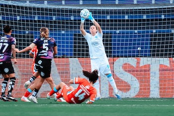 Una portera con uniforme blanco atrapa el balón en el aire frente al arco mientras varias futbolistas disputan la jugada