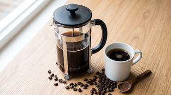 Cafetera de émbolo con café negro, taza blanca llena, granos de café dispersos y una cuchara pequeña sobre una mesa de madera clara.