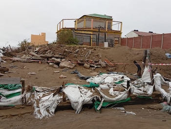 Vista de una playa con arena y abundante basura, escombros y sacos de arena rotos en primer plano. Al fondo, un edificio de dos pisos deteriorado