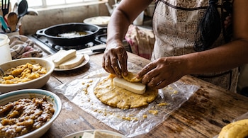 Manos doblando una masa amarilla sobre un trozo de queso blanco en una mesa de madera con recipientes y una sartén al fondo.