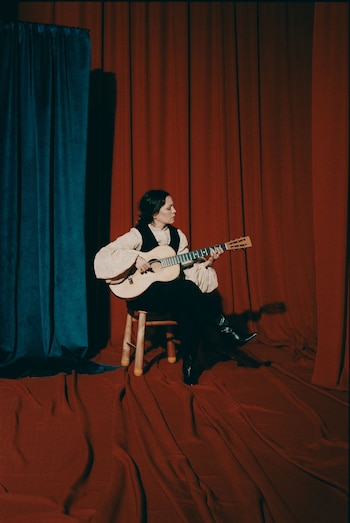 Natalia Lafourcade sentada en un taburete de madera, tocando una guitarra acústica frente a cortinas de teatro rojas y azules