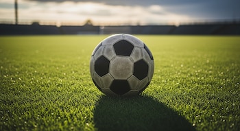 Un balón de fútbol blanco y negro sobre un campo de césped verde. Al fondo se ven las gradas de un estadio bajo un cielo parcialmente nublado.