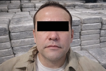 Primer plano de un hombre con los ojos censurados, vistiendo una camisa beige, con numerosas pilas de paquetes rectangulares de droga en el fondo.