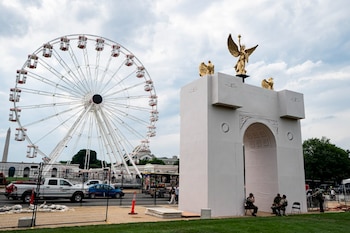 Una rueda de la fortuna blanca, un arco conmemorativo blanco con tres esculturas doradas, vehículos, personas, vallas y el cielo con nubes