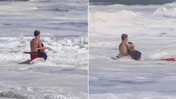 Un adulto y un niño están en el mar. El adulto viste bañador rojo y sujeta al niño. Una boya de rescate negra y roja flota cerca. Hay olas y espuma