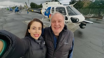 Un hombre y una mujer sonrientes posan en primer plano con un helicóptero blanco de noticias y una pista asfaltada de fondo