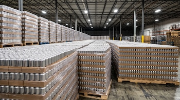 Palés de madera cargados con latas metálicas sin etiquetar y envueltas en plástico en un almacén.