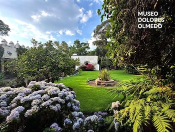 Un jardín con césped verde, arbustos de hortensias lilas y blancas, helechos, árboles, y un edificio blanco con almenas al fondo bajo un cielo azul con nubes