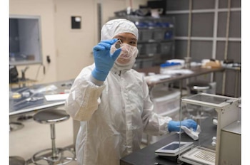 Una persona con traje blanco, mascarilla y guantes sostiene un pequeño disco metálico junto a una balanza de precisión en un laboratorio