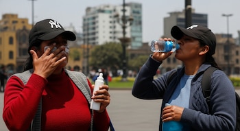 Dos mujeres al aire libre; una aplica crema blanca en su cara y sostiene un envase, la otra bebe agua de una botella. Ambas usan gorras.