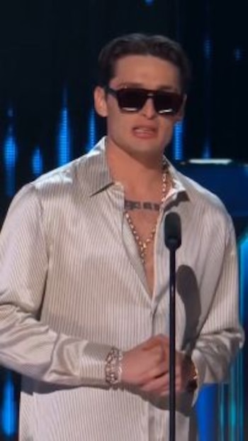Un hombre con gafas de sol oscuras y camisa clara de rayas se encuentra frente a un micrófono de pie