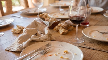 Vista de cerca de una mesa de madera con platos blancos sucios, cubiertos, migas de pan y varias copas de vino tinto, una de ellas en primer plano.
