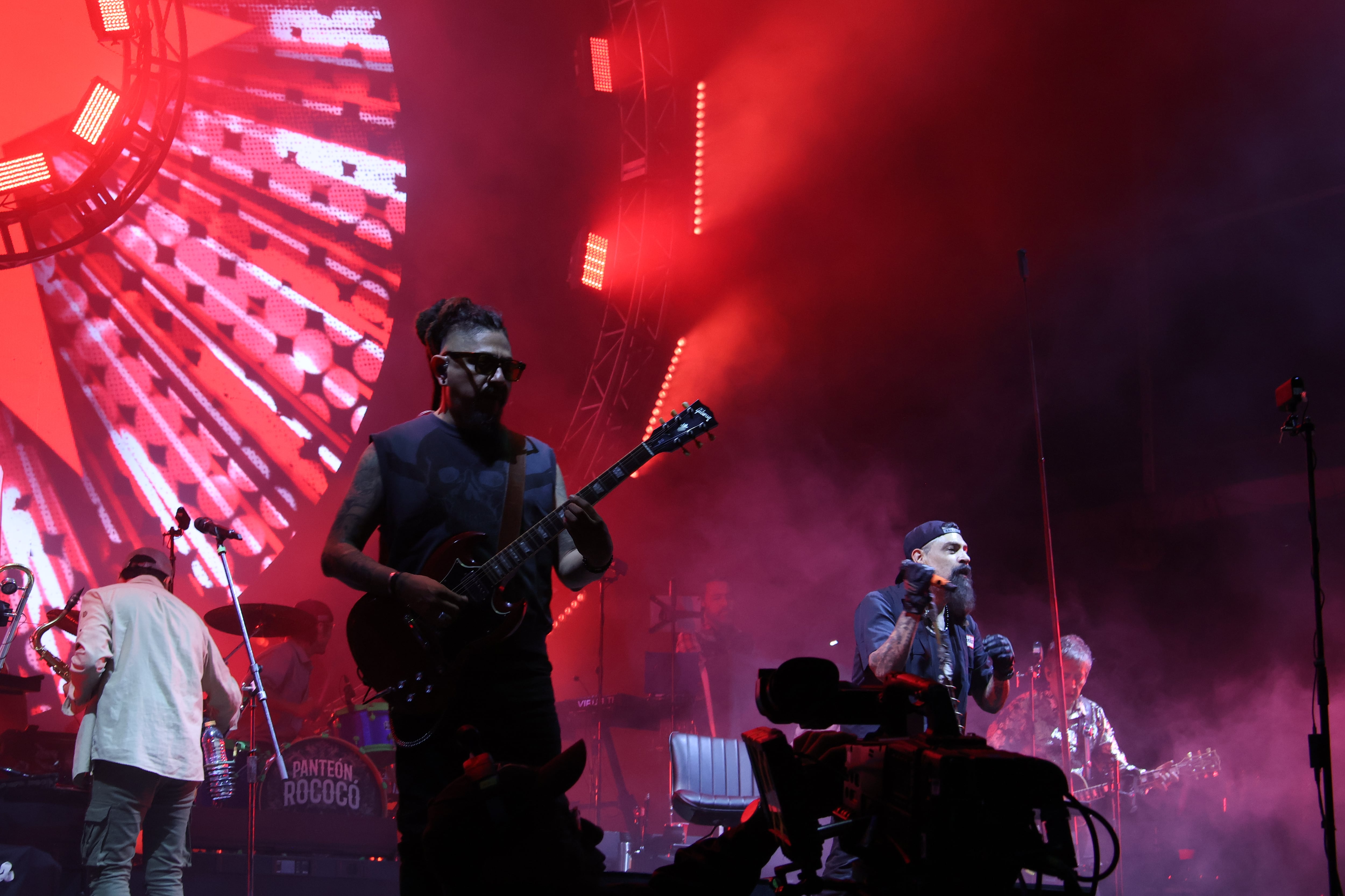 Un guitarrista de la banda Panteón Rococó toca con pasión bajo luces rojas durante un enérgico concierto de rock. (Selene Miranda/ Infobae México )