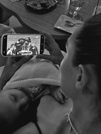 Fotografía en blanco y negro de una mujer que sostiene a un bebé dormido mientras observa un partido de fútbol en la pantalla de un teléfono