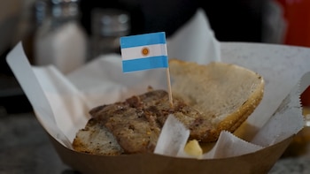 Pan tostado con trozo de carne en envase de cartón y papel, con bandera de Argentina insertada en la comida