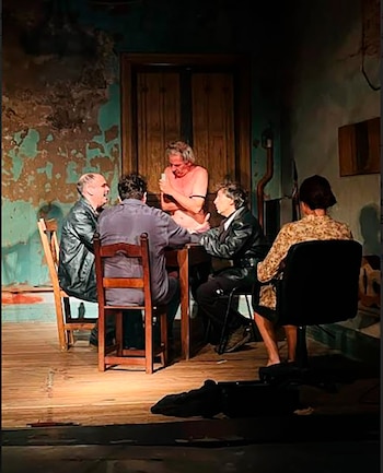 Cinco personas, cuatro hombres y una mujer, sentadas alrededor de una mesa de madera en un escenario con una pared texturizada y una puerta antigua de madera al fondo