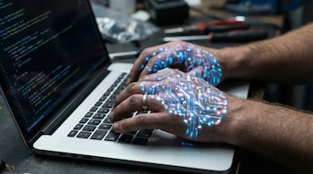 Primer plano de las manos de un ingeniero escribiendo en un teclado de laptop. Un patrón luminoso de circuito digital se proyecta sobre sus manos y el teclado.