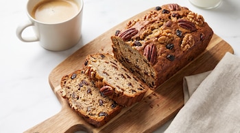 Pan de plátano rebanado con nueces y pasas sobre una tabla de madera, acompañado por una taza de café.