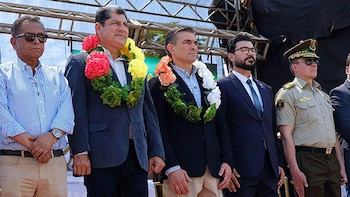 Cinco hombres de pie. Tres con guirnaldas florales. Un hombre de uniforme militar y gorra. Otro con traje y corbata. Fondo con estructuras metálicas