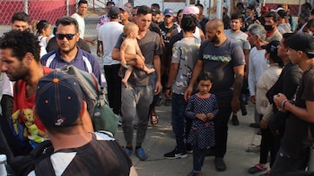 Una multitud de personas, incluyendo adultos y niños, se congrega en un espacio exterior. Un hombre lleva un bebé y una niña pequeña está cerca