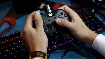 Primer plano de unas manos sujetando un controlador de videojuegos Xbox negro con los botones iluminados, sobre un teclado de gaming con luces RGB