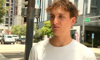 Un joven de cabello castaño claro y camiseta blanca se encuentra en una calle urbana con edificios, coches, semáforos y árboles al fondo