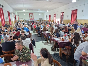 Vista interior de un amplio salón con numerosas personas sentadas en mesas de madera, algunas aplaudiendo, y un estandarte rojo con un escudo en la pared de fondo