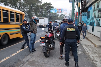 Agentes de policía inspeccionan a motociclistas en una calle. Un oficial revisa el casco de un conductor junto a su moto, con un autobús y cartel al fondo