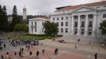 Vista aérea do campus universitário com estudantes ambulantes e edifícios imponentes, incluindo a torre alta à esquerda, sob um céu branco