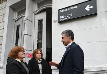 Un hombre de mediana edad conversa con dos mujeres en la calle Bahía Blanca. Detrás, un cartel negro indica 'Bahía Blanca 300-200 Comuna 10' y un edificio blanco