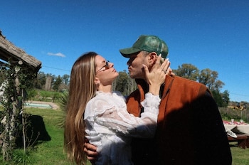 Una mujer con blusa blanca y un hombre con gorra verde y chaleco marrón junta sus rostros en un campo con cielo despejado