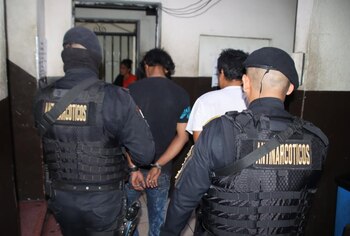 Dos agentes de uniforme negro con chalecos 'Antinarcóticos' escoltan a tres hombres, uno de ellos con esposas
