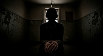 Una persona sentada de espaldas a una ventana, con el rostro en sombra, en una habitación oscura de paredes descascaradas. Una bombilla cuelga del techo.