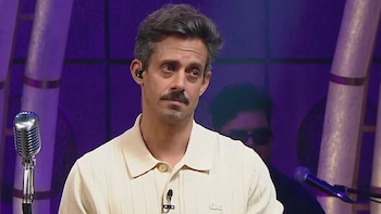 Un hombre con bigote, camisa polo beige y audífono, mira pensativo. Hay un micrófono antiguo y un fondo púrpura con columnas verticales