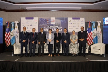 Nueve personas de pie en un escenario. Telón de fondo con banderas de Estados Unidos y Guatemala. Banderas en asta de Estados Unidos, Guatemala y AmCham