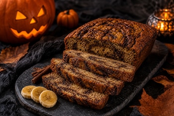 Pan de plátano en rebanadas sobre una tabla negra, junto a rodajas de plátano, ramas de canela y calabazas de Halloween iluminadas.