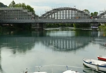 Vista de un puente de arco y celosía cubierto de andamios sobre un río, con vegetación en la orilla distante y botes blancos en primer plano