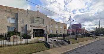 Exterior del Mason Temple, un edificio de piedra con cruz en el techo y estatua. Verja negra, césped, acera y un letrero de 'Church of God in Christ' al fondo visible