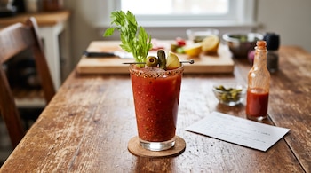 Vaso con bebida roja decorado con apio, aceituna y limón sobre un posavasos junto a una botella de salsa picante y una tarjeta escrita.