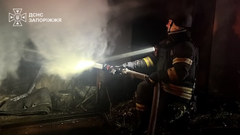Un bombero equipado con casco y linterna arroja agua con una manguera en una estructura oscura llena de humo