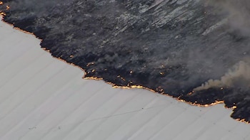 Una vista aérea muestra una línea de fuego y humo denso en el techo gris oscuro de una estructura, con la parte inferior del techo blanca y brillante