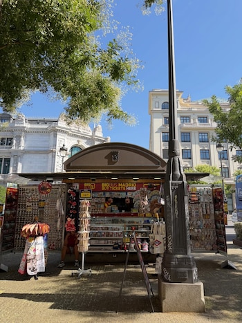 Puesto callejero con techo marrón y carteles de 'Big Bus Madrid', exhibiendo souvenirs y pulseras. Un farol negro, árboles verdes y edificios de fachada blanca
