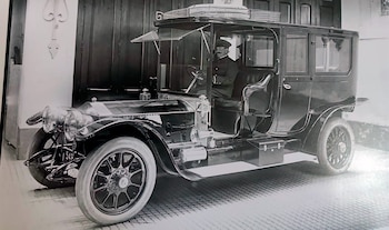 Un automóvil antiguo de color oscuro con un chófer uniformado al volante frente a una pared de madera