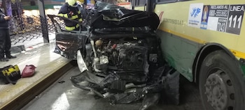 Automóvil negro con frontal destrozado contra el lateral de un autobús. Un bombero, una persona de oscuro, una valla metálica y escombros en el suelo